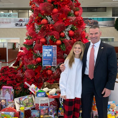 Ella beside a decorated Christmas tree and a Pediatric Cancer Toy Drive sign