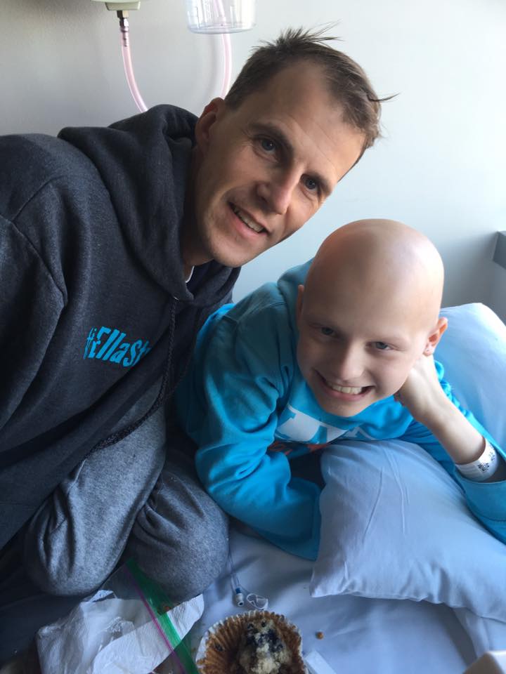 Ella smiling with her dad, in an EllaStrong sweatshirt, at her hospital bedside