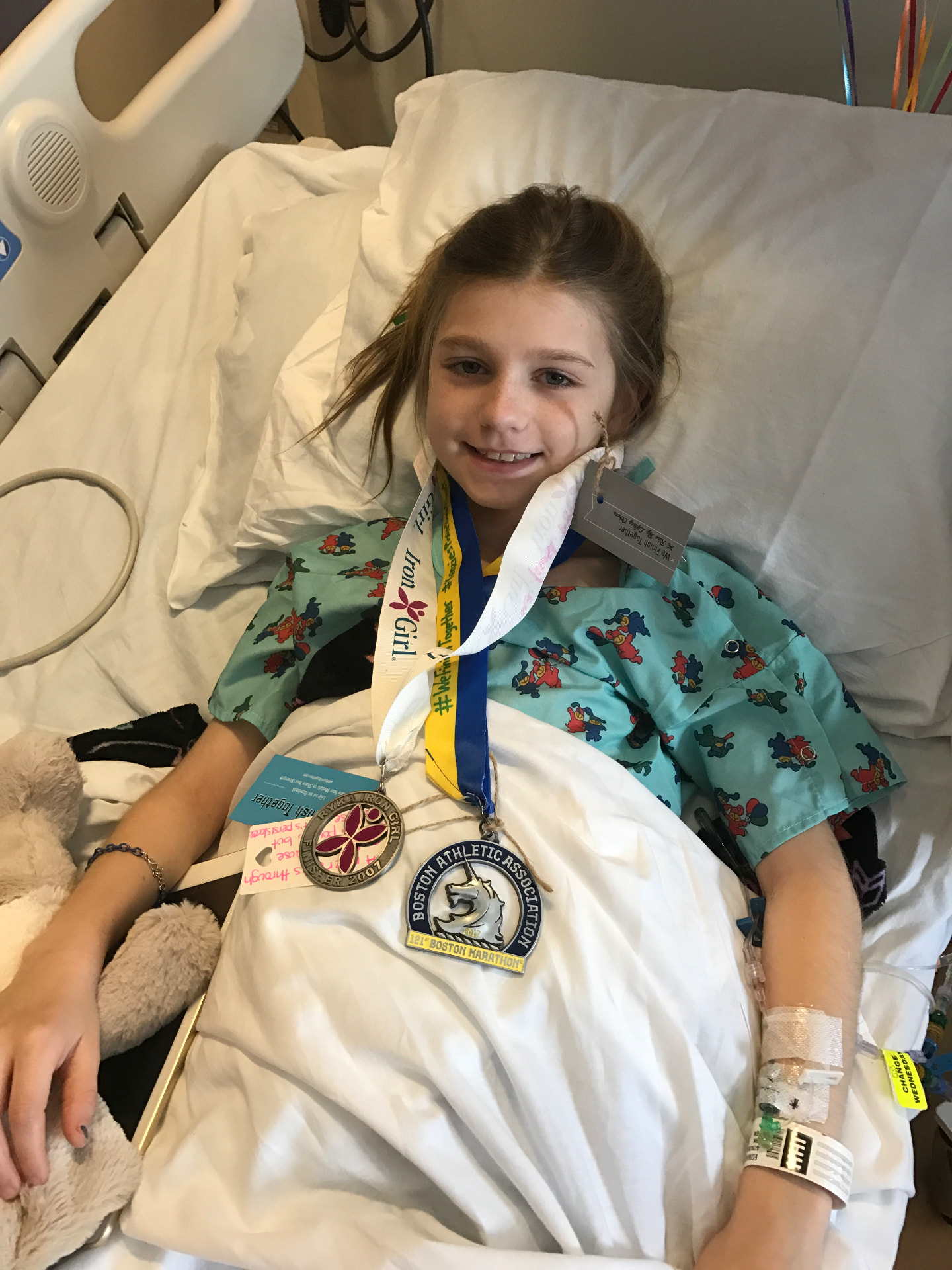 Ella in her hospital bed wearing Boston Marathon and Iron Girl medals given to her by runners