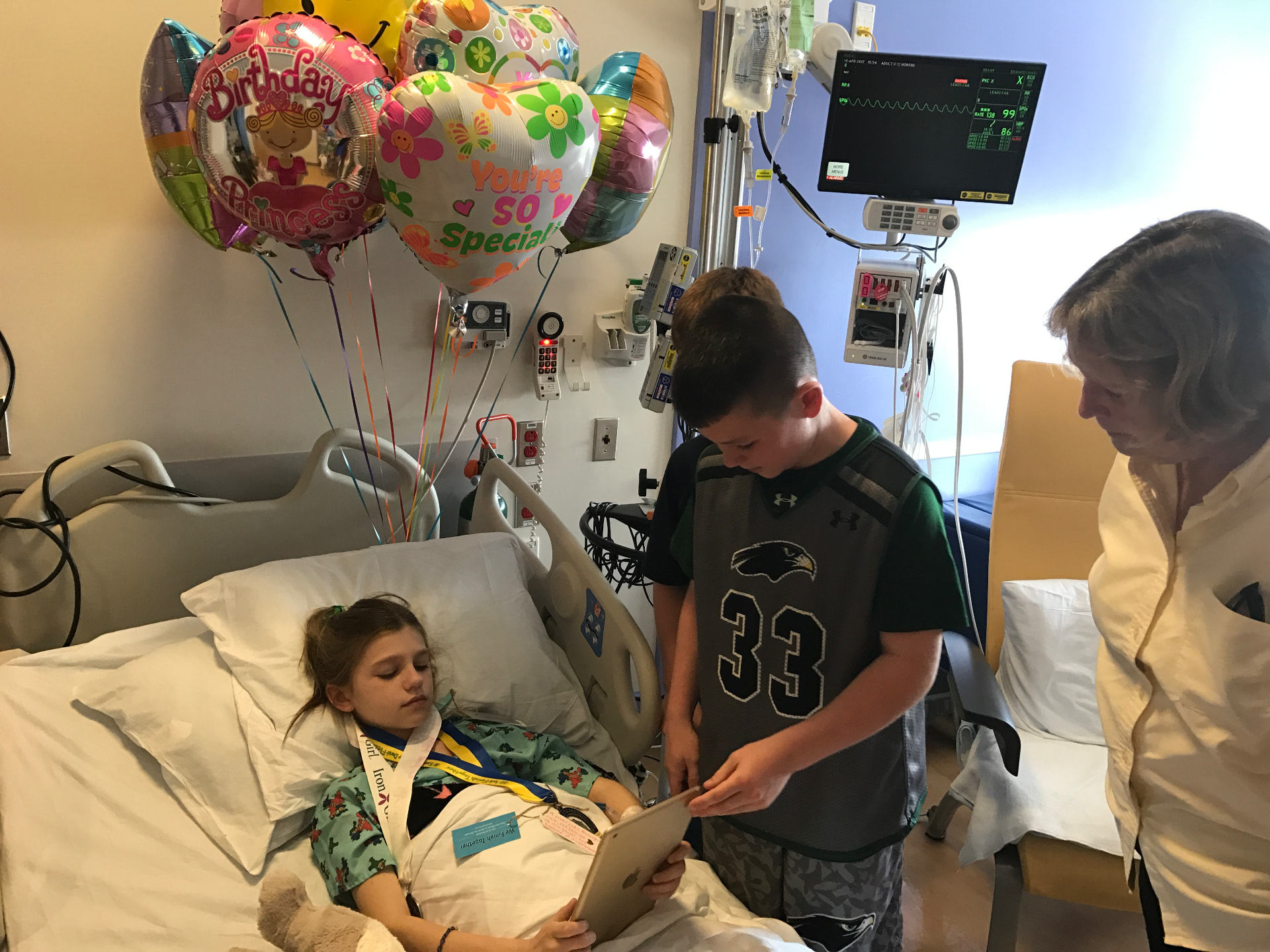 Ella in a hospital bed on her birthday surrounded by balloons as her brother shows her a tablet